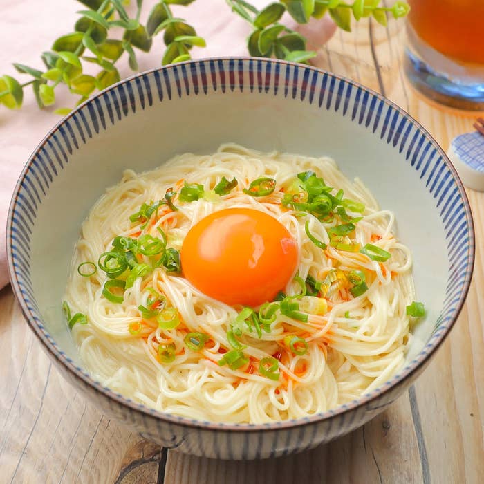 器に盛られた素麺の上に生卵とネギが載っている。背景に観葉植物が飾られている。