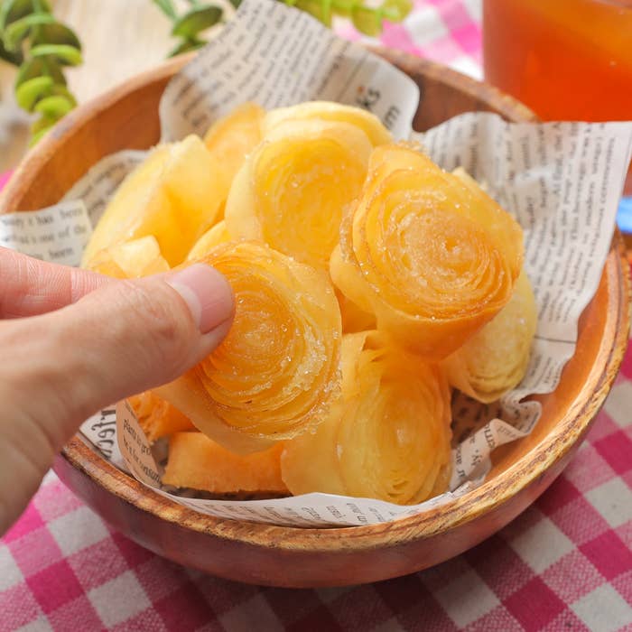 手で持っている揚げたスイーツが木のボウルに入っている。背景に植物と飲み物がある。