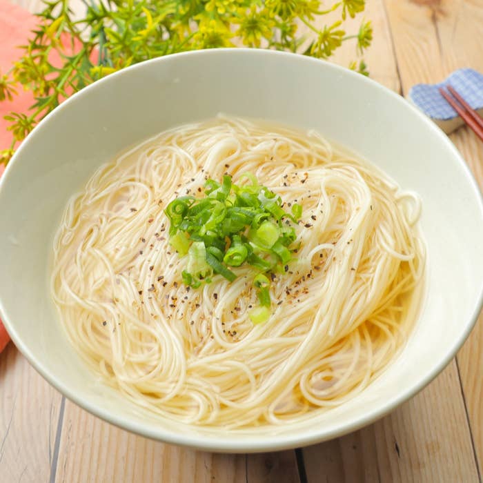 白い器に入ったそうめん、上に刻みネギとこしょうが振りかけられています。背景に花があります。