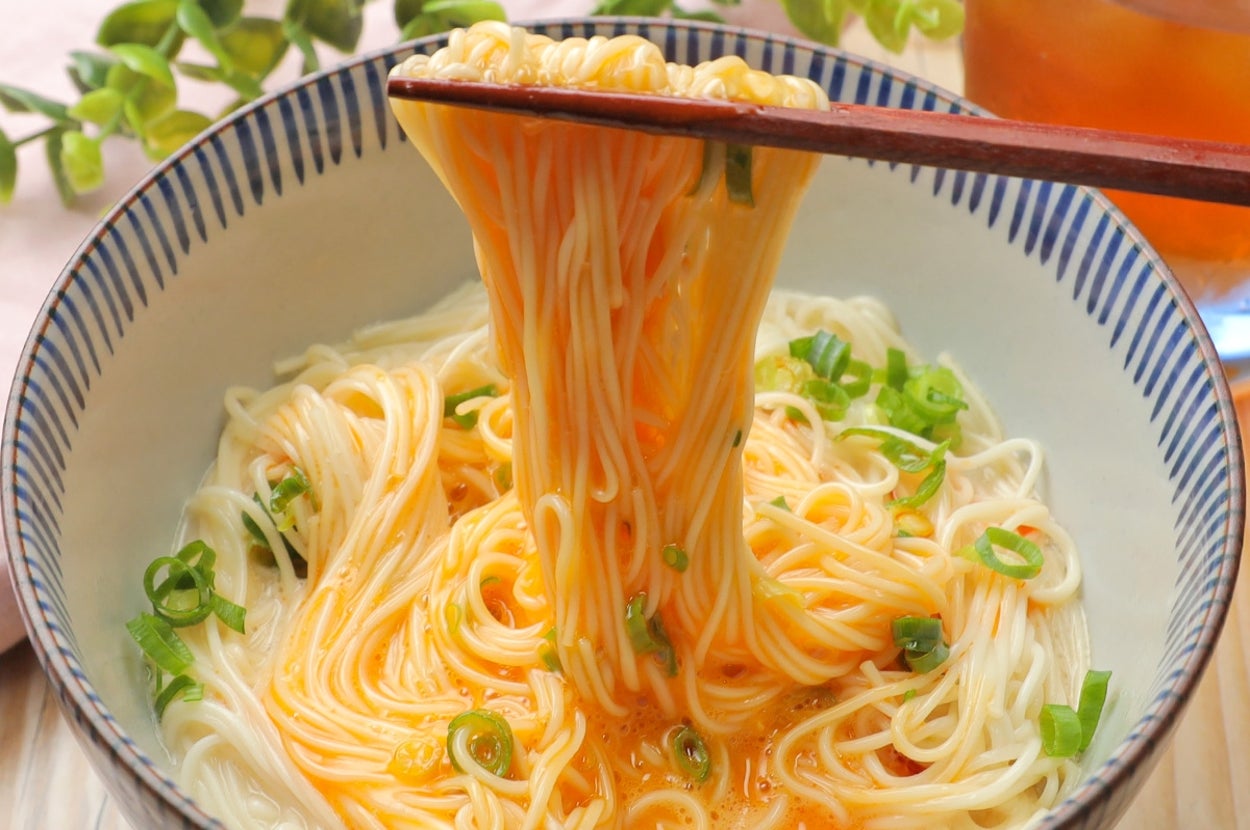 器に盛られた素麺から箸で持ち上げた麺。トッピングにネギが散らされています。背景に植物の装飾。