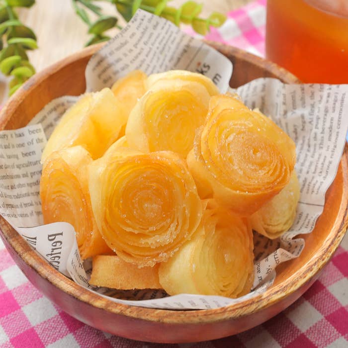 木製のボウルに盛られた、バラの形をした美しい焼き菓子。背景に飲み物と緑の葉。