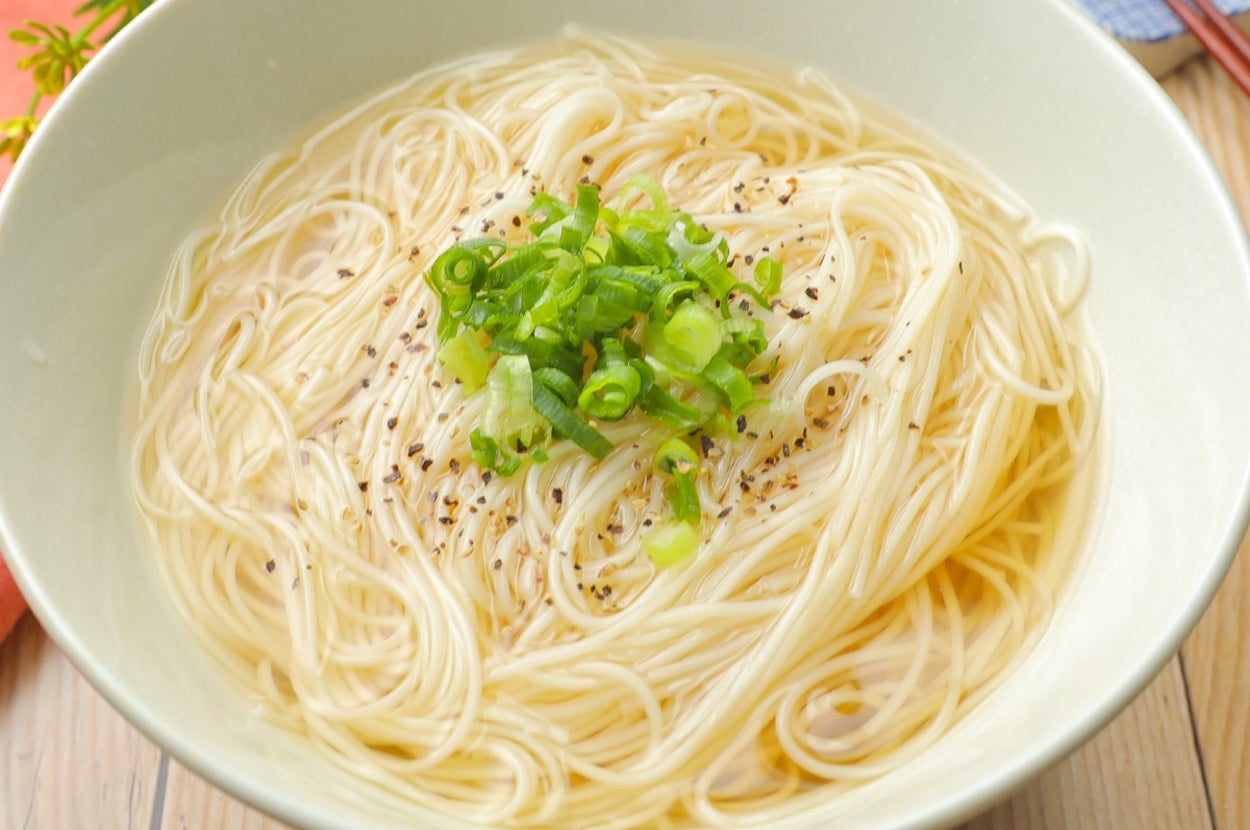 白いボウルに盛られた素麺。上に刻みネギと黒コショウが散らされている。背景に黄色の花が見える。