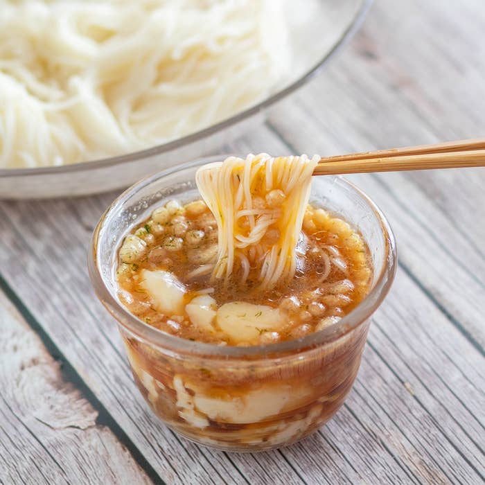 テーブル上の素麺を箸でつまみ、だしに浸す様子。背景に素麺の盛られたボウルが見える。