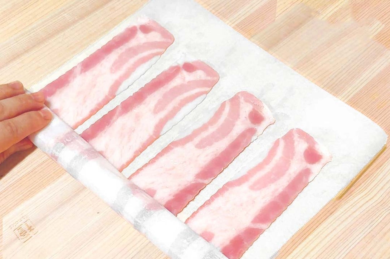 手がキッチンペーパーに並べられた薄切りベーコンを持ち上げようとしている様子。
