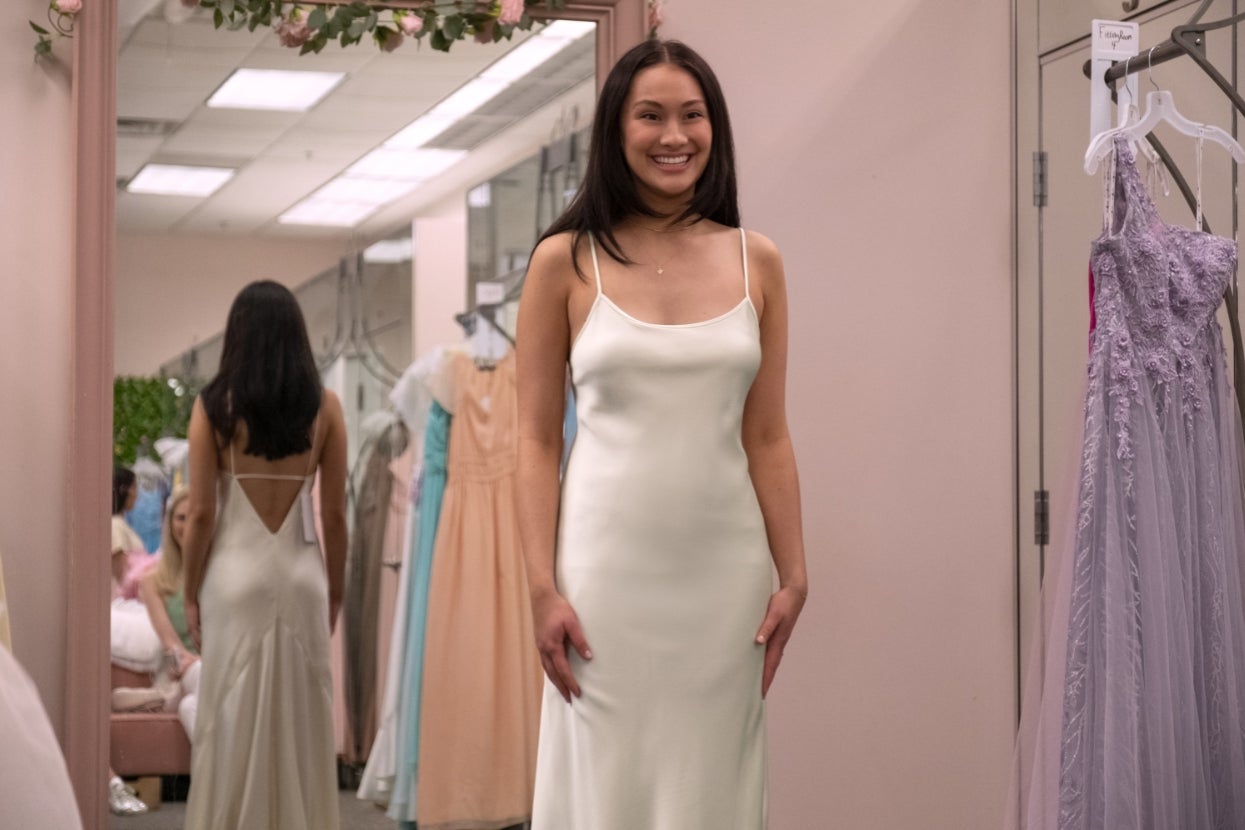 Lola Tung as Belly stands smiling in a bridal shop wearing a sleek slip-style dress, surrounded by other dresses on display