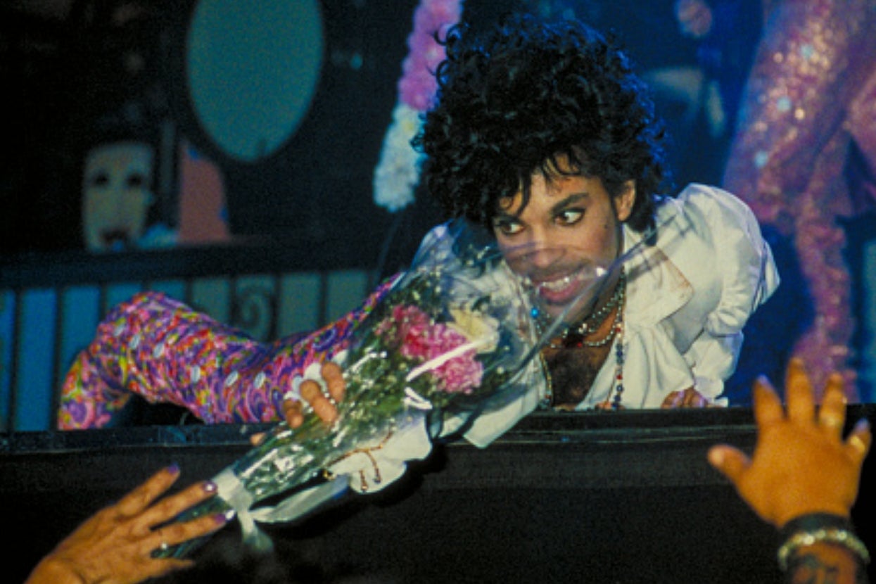 A performer onstage in an ornate shirt and colorful pants, holding flowers while engaging with fans during a concert
