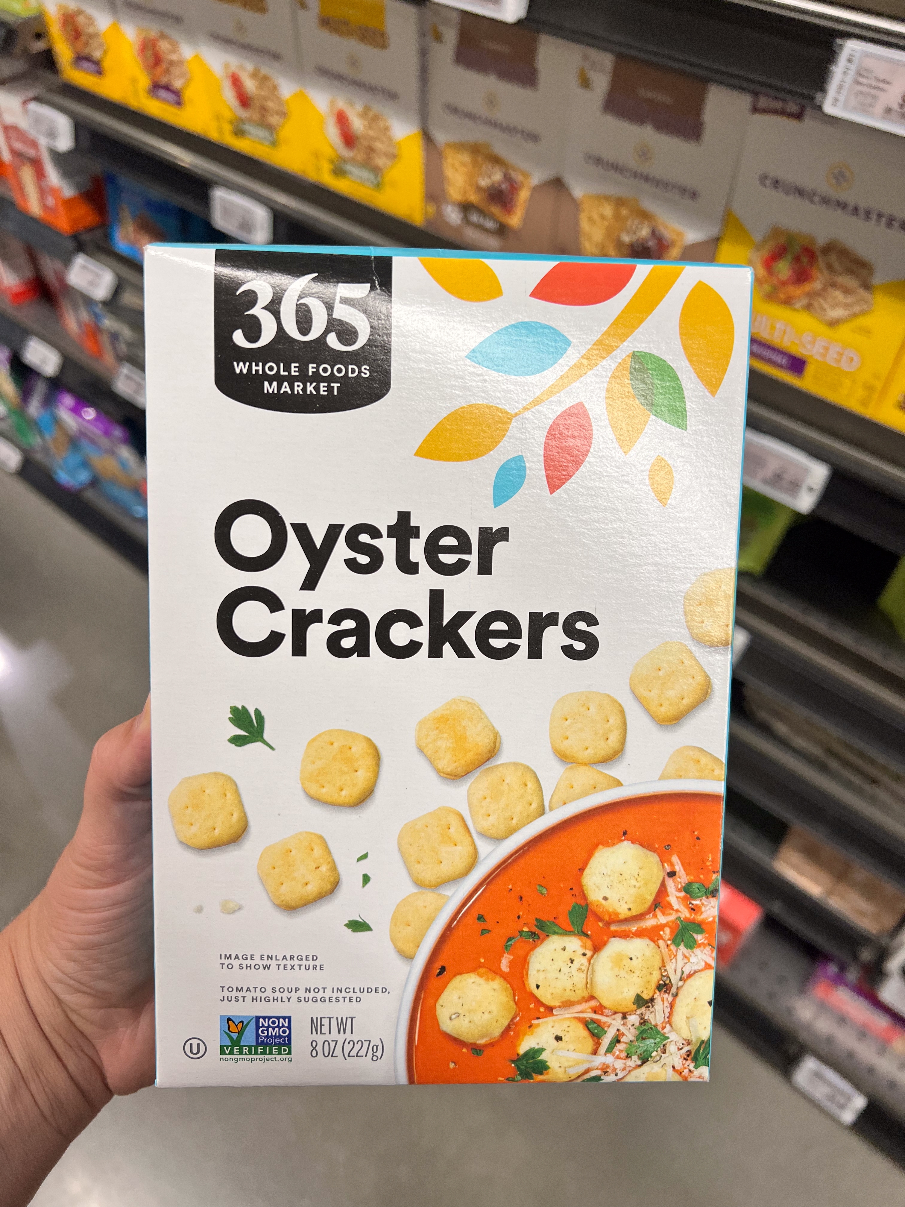 A hand holding a box of 365 Whole Foods Market Oyster Crackers in a grocery aisle