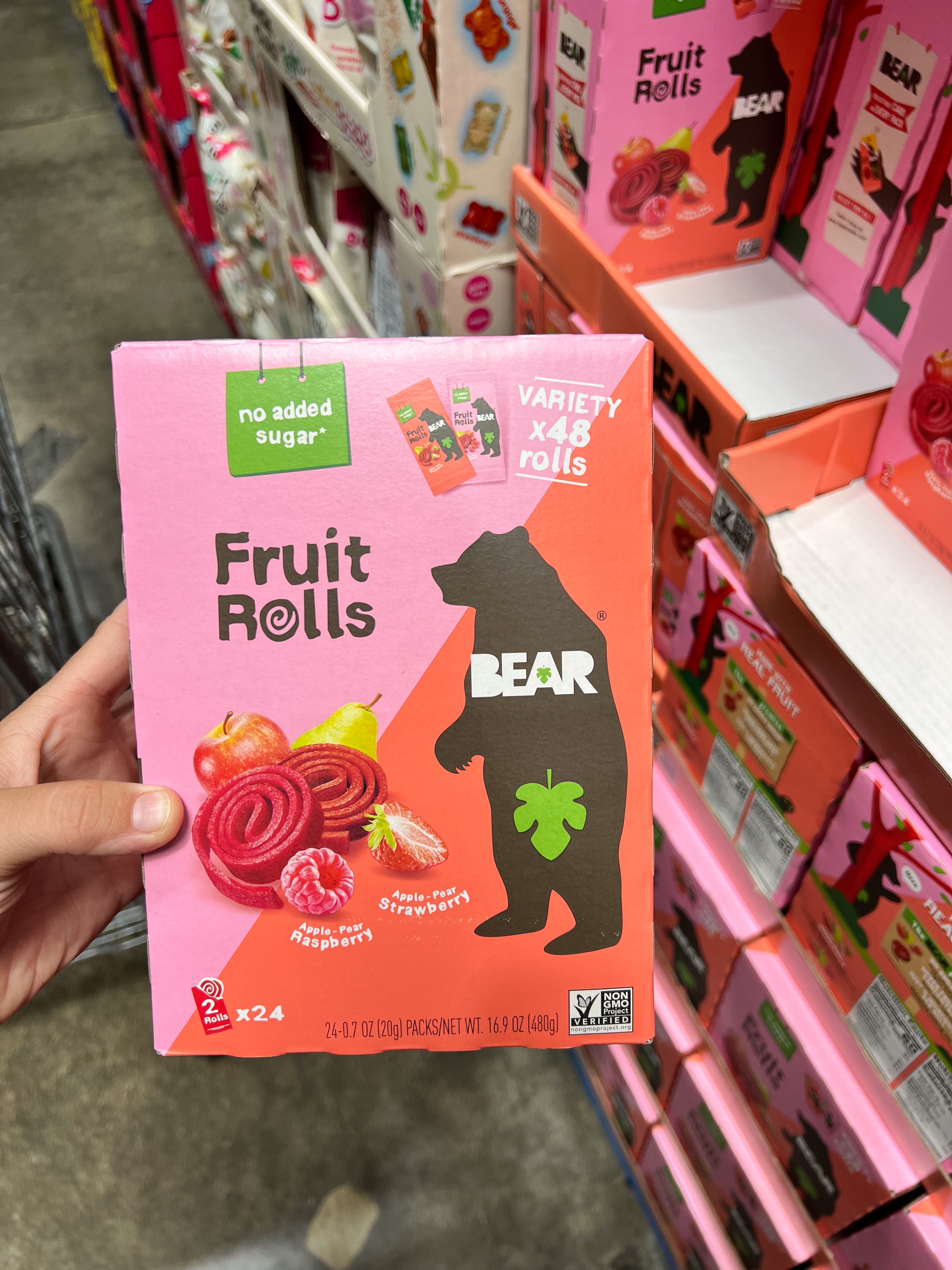 A person holds a box of BEAR fruit rolls from a store display. The box highlights "no added sugar" and contains images of fruit