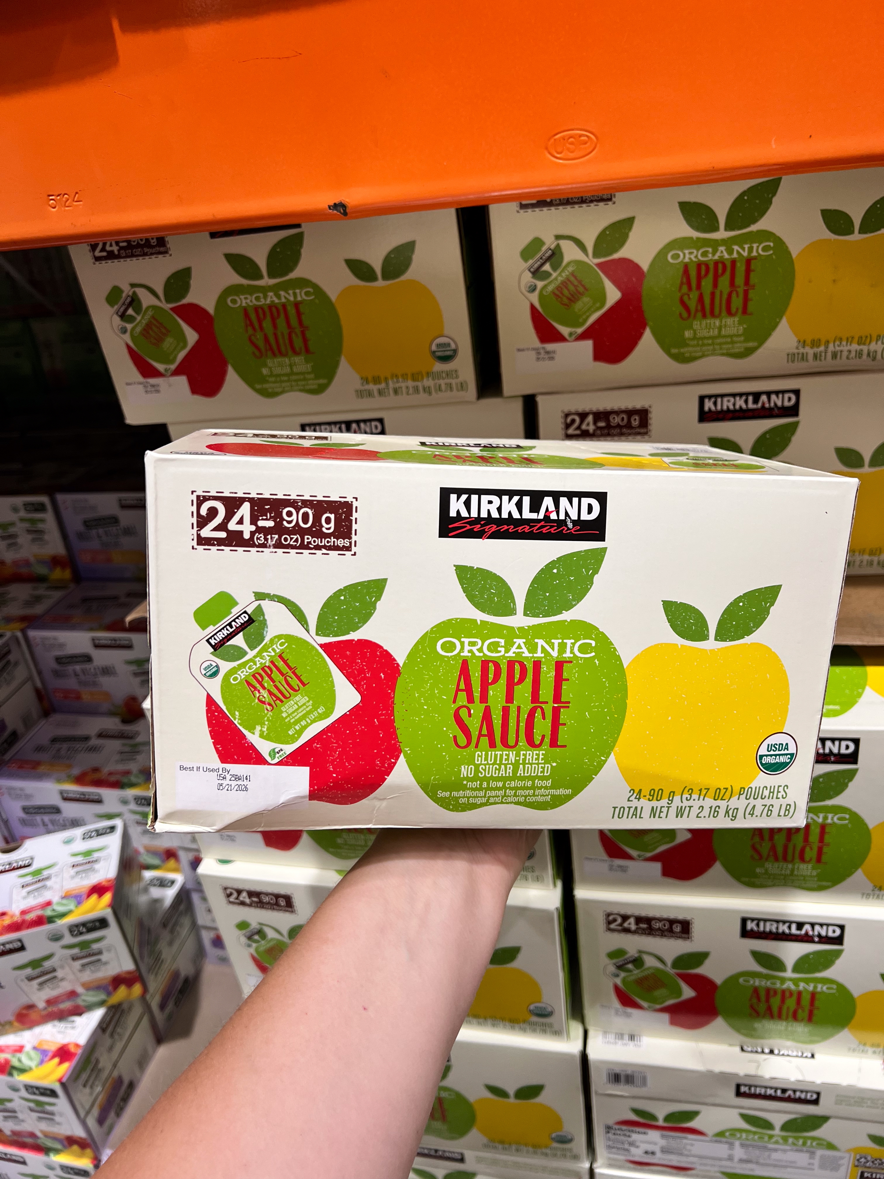 Person holding a box of organic apple sauce pouches in a store, with shelves of similar products in the background