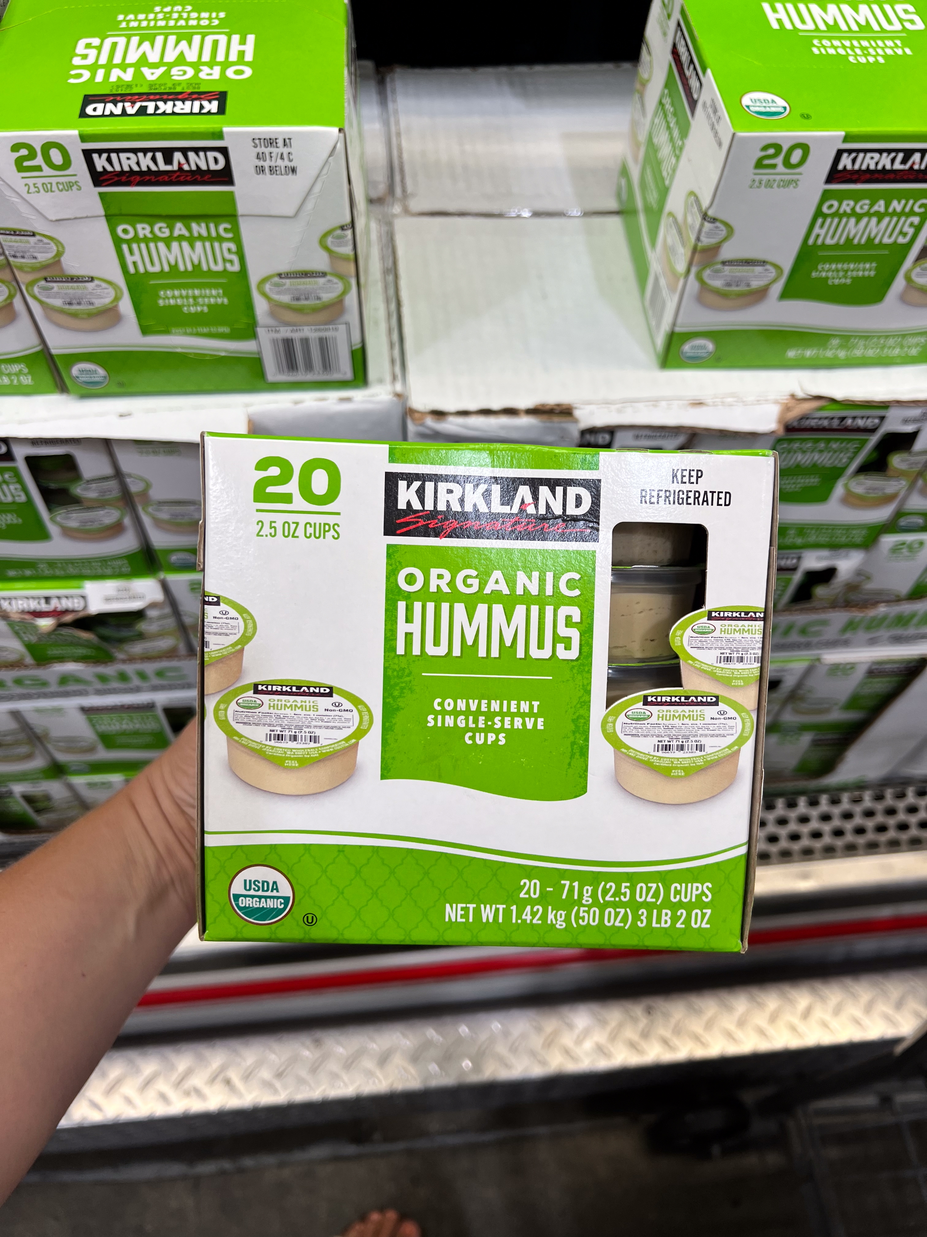 Hand holding a box of Kirkland Organic Hummus, 20 single-serve cups, at a grocery store display
