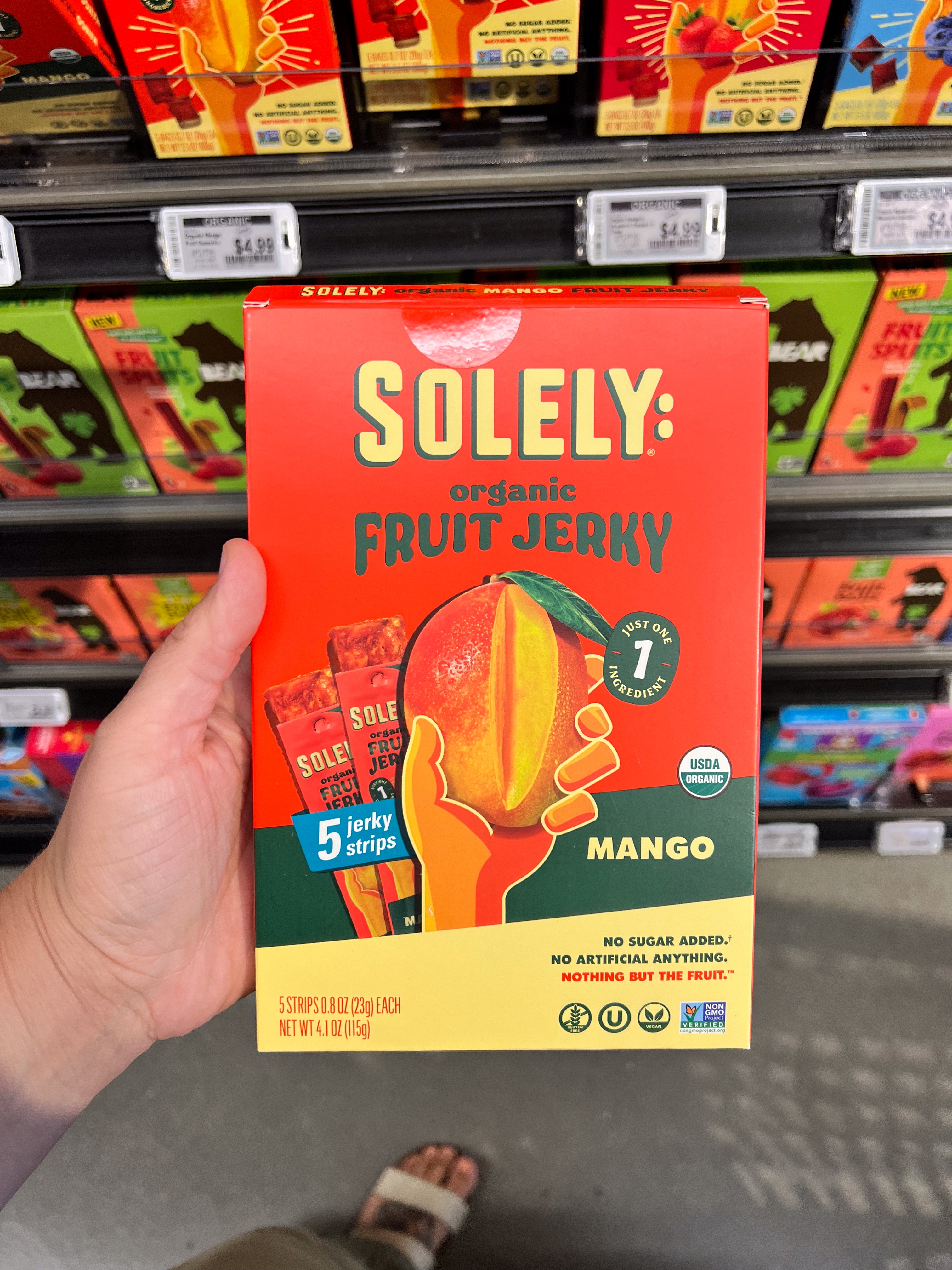 Hand holding a box of Solely Organic Fruit Jerky, Mango flavor, in a store aisle. Packaging highlights no added sugar and USDA organic certification