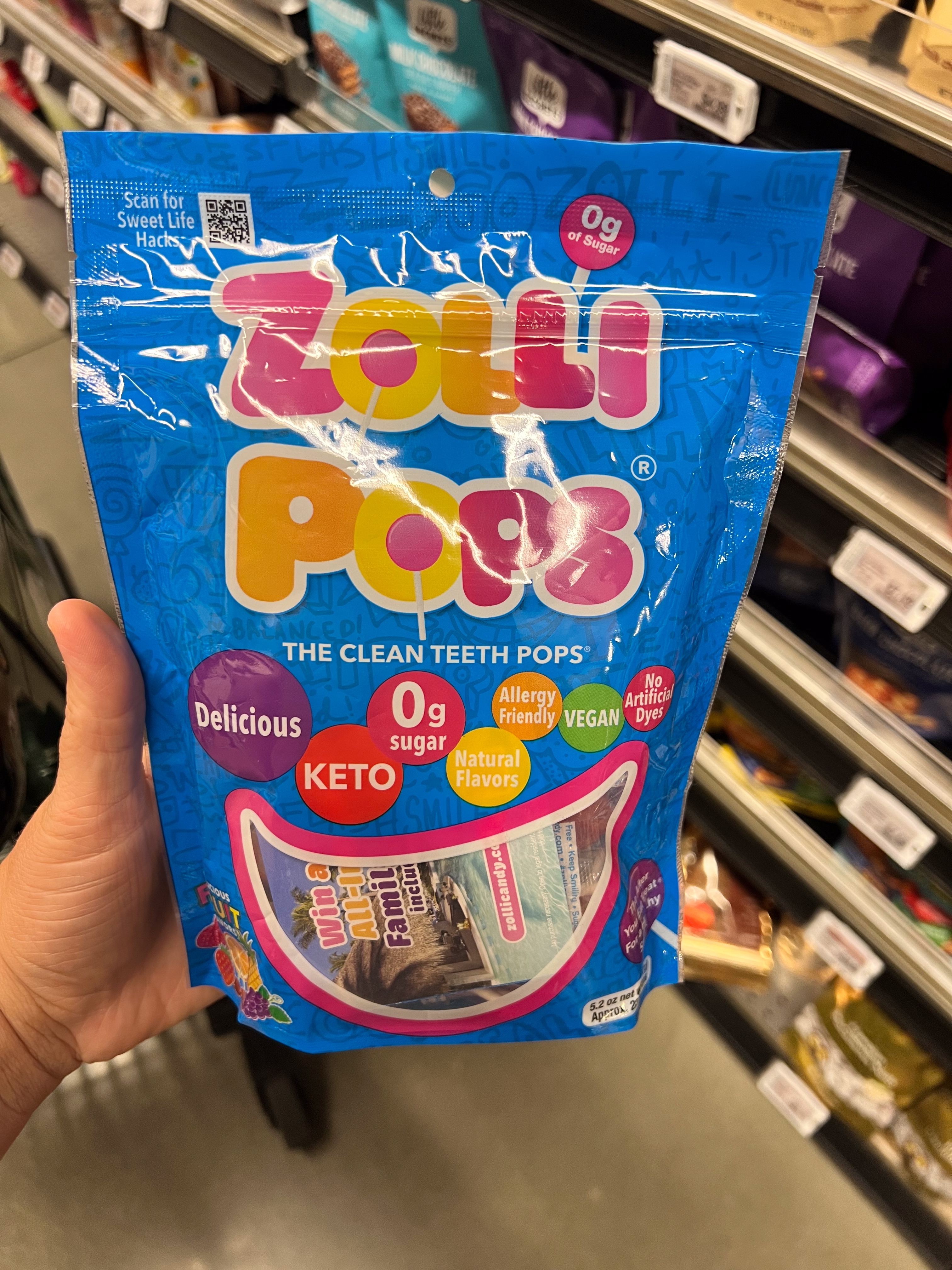 Bag of Zolli Pops held in a store aisle. Package highlights include "clean teeth pops," "0g sugar," "KETO," "vegan," and "natural flavors."
