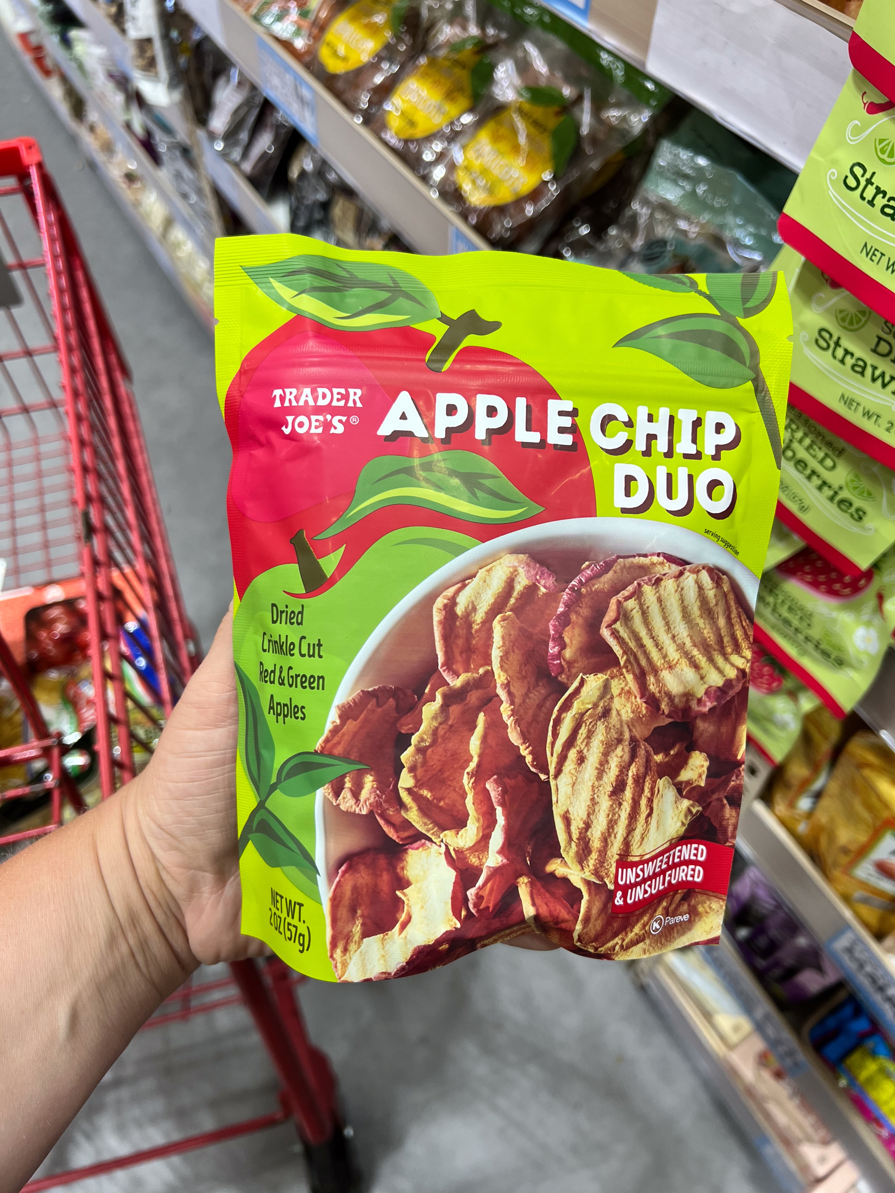 A hand holds a bag of Trader Joe's Apple Chip Duo, featuring dried apple chips. The background shows a grocery shelf with various snacks