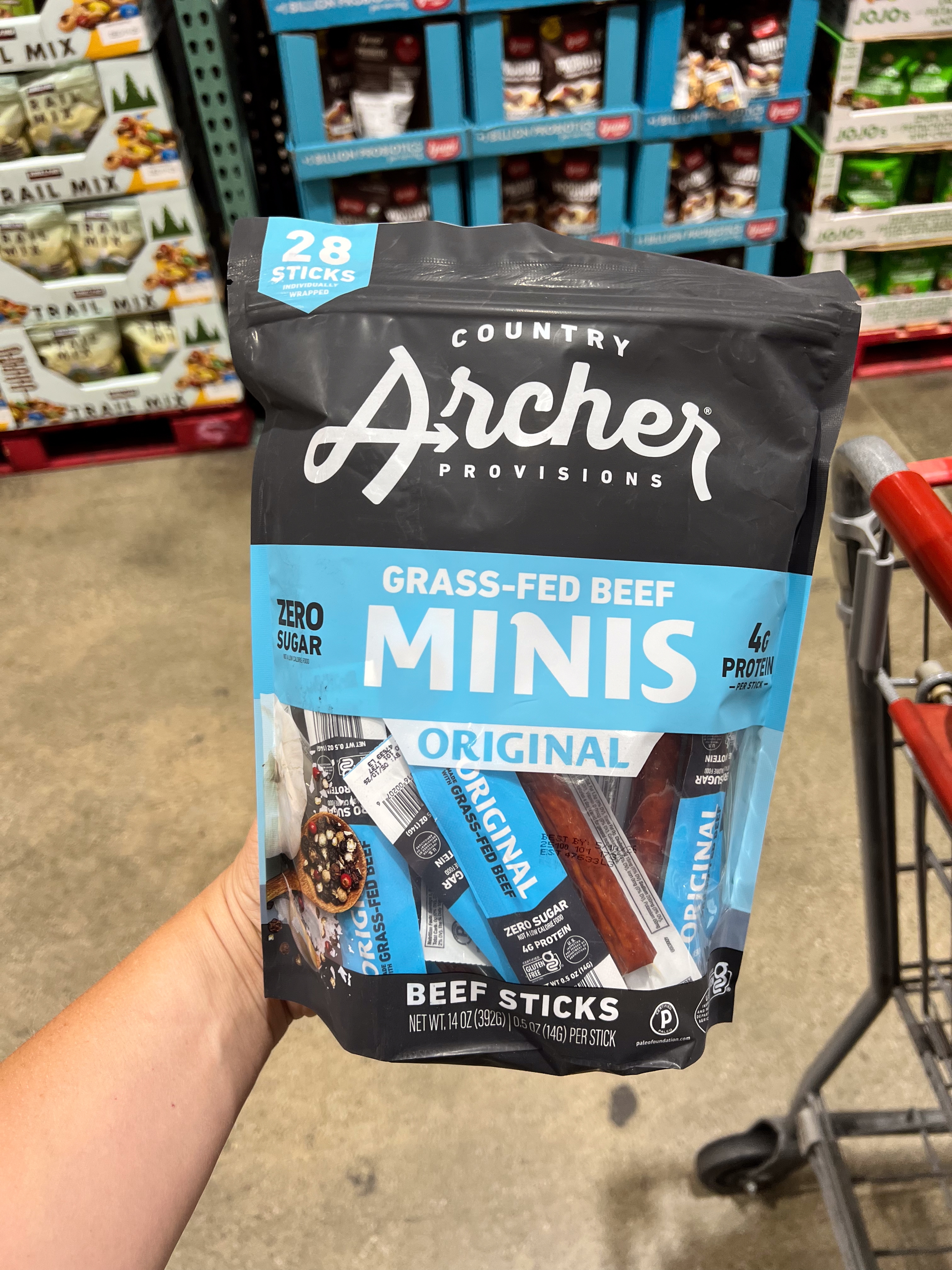 Person holding a package of Country Archer grass-fed beef minis in a store aisle, with a shopping cart partially visible
