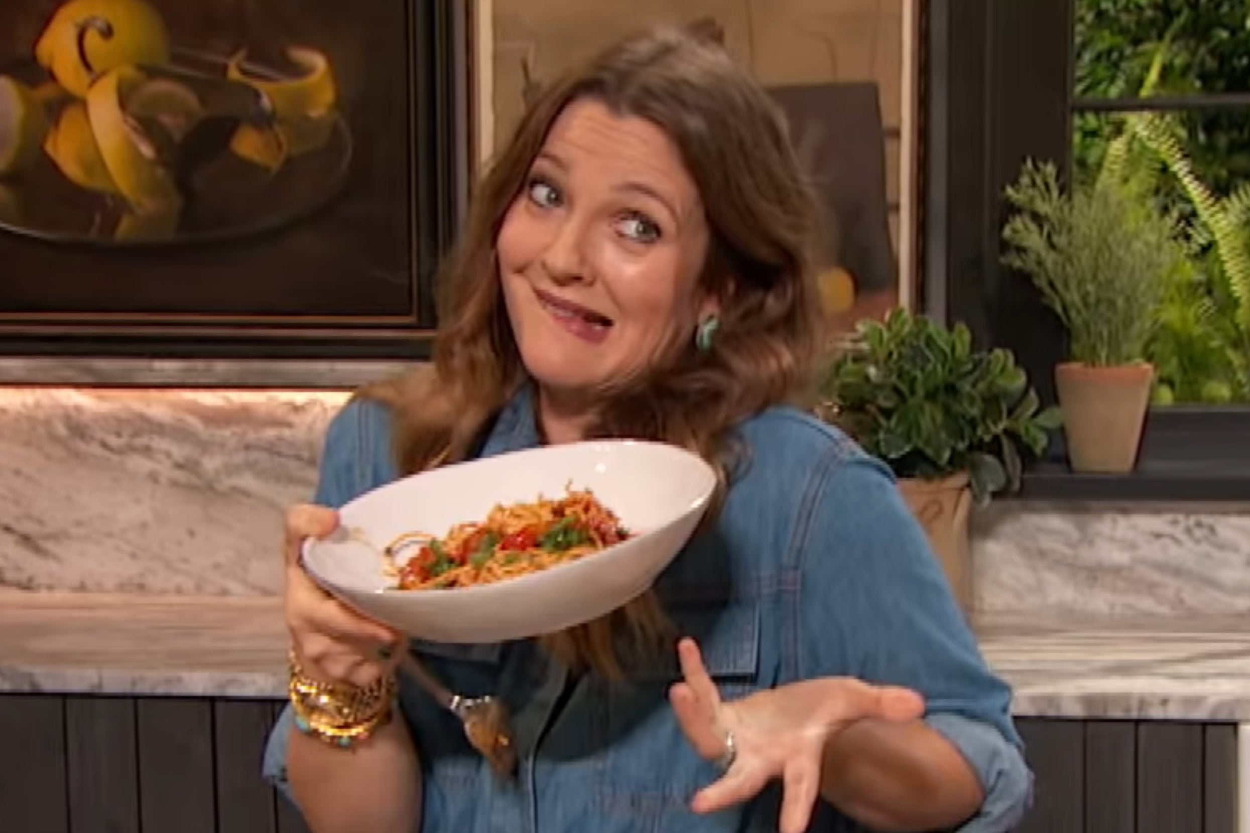 Drew Barrymore in a denim shirt holds a bowl of pasta, making a playful shrugging gesture, in a kitchen setting