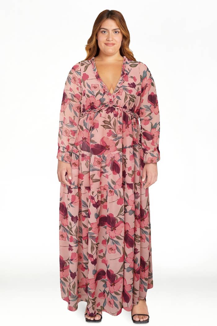 Person wearing a floral maxi formal with agelong sleeves and a tied waist, lasting against a plain backdrop.