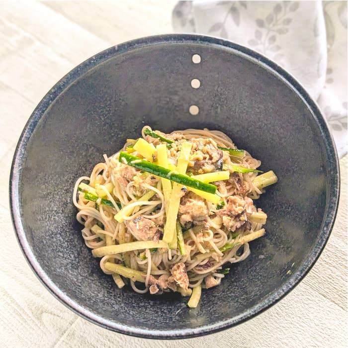 黒いボウルに盛られたそうめん、細切りの野菜とサバがトッピングされた一皿の料理。白い布が横に置かれている。