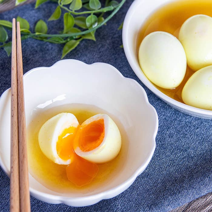 割った味玉が皿に盛られ、つけ汁に浸っています。隣にはつけ汁入りの味玉2つが見えます。背景に緑の装飾があります。