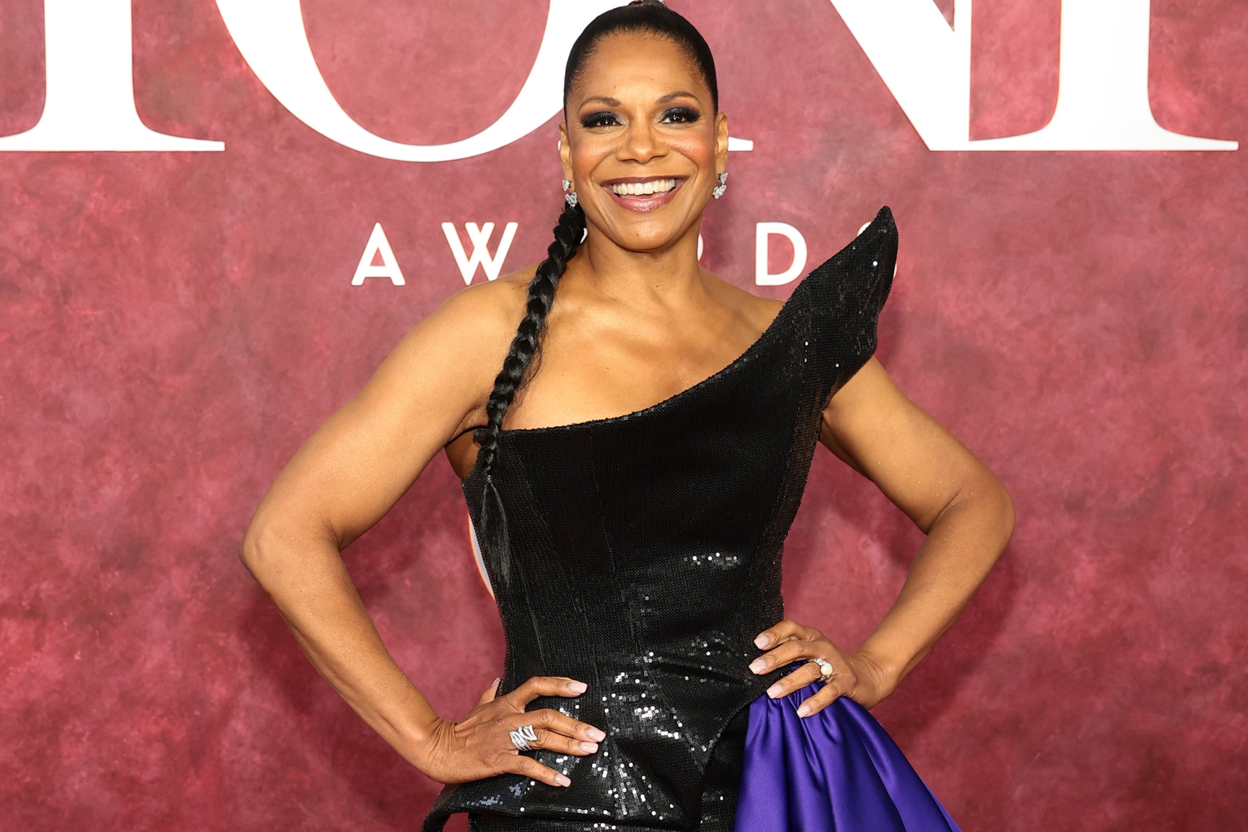 Celebrity poses on red carpet in a sleek, black one-shoulder gown with a dramatic side train at the Tony Awards