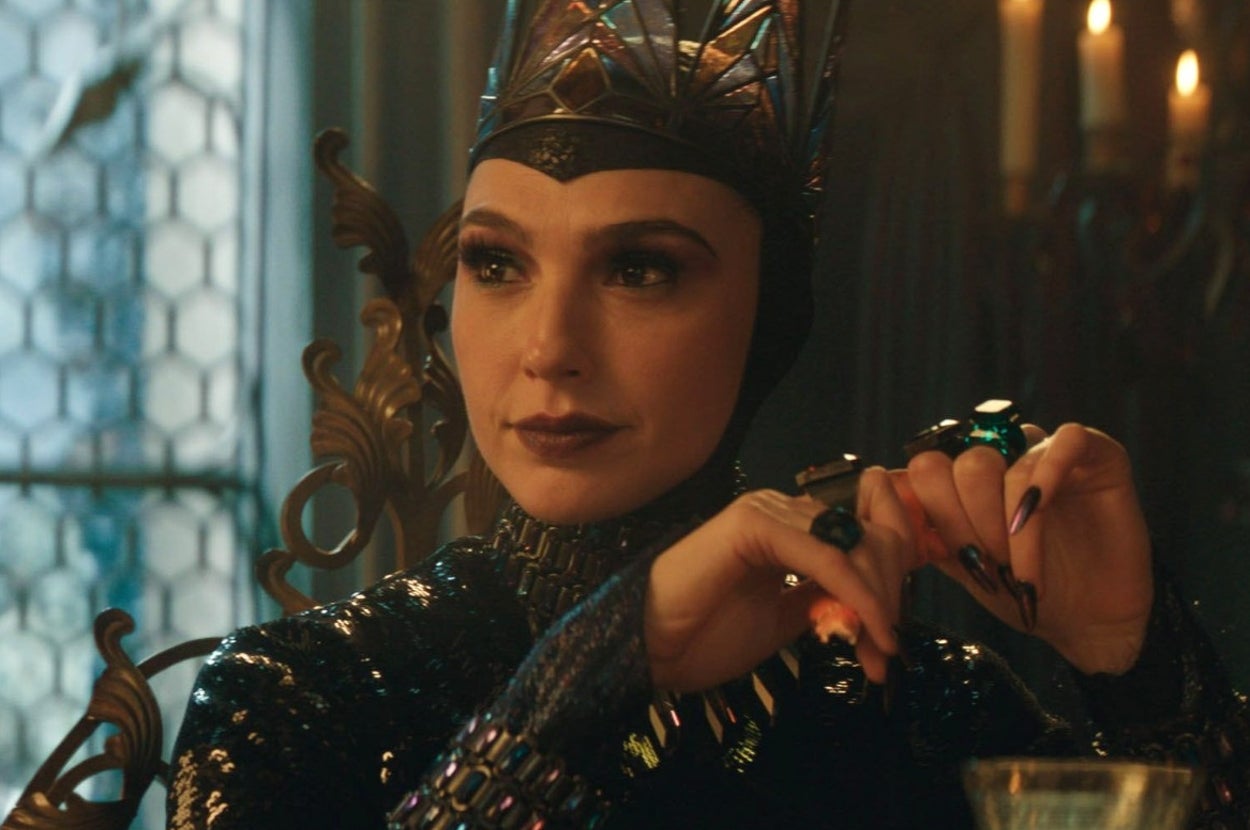 Two people in an ornate room, one in a detailed, dark fantasy costume with a crown, seated at a table with cards and ornate items