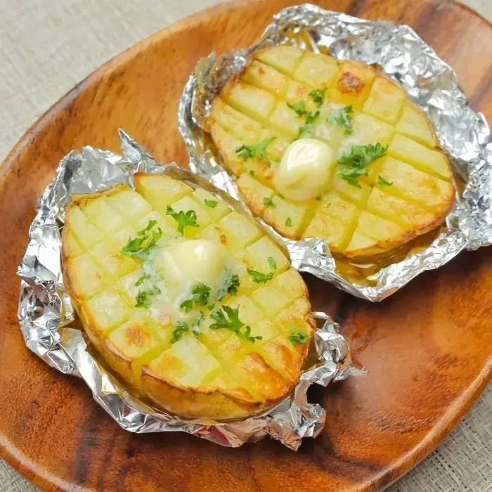 ホイルに包まれた焼きジャガイモが2つ、バターとパセリが添えられて木製の皿に乗っている。