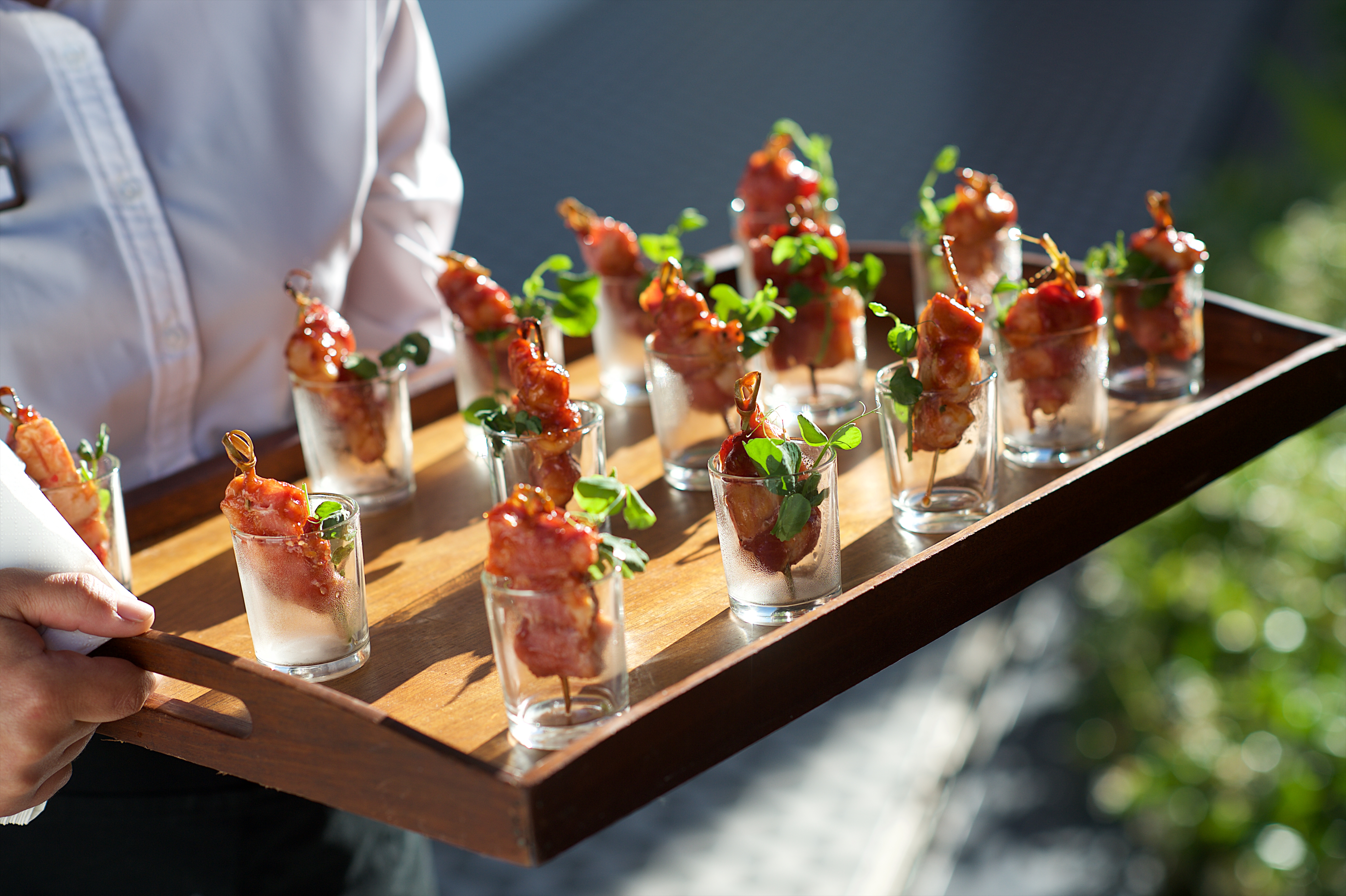 Tray of appetizers with bacon skewers in shot glasses, garnished with green herbs, held by a person in a white shirt