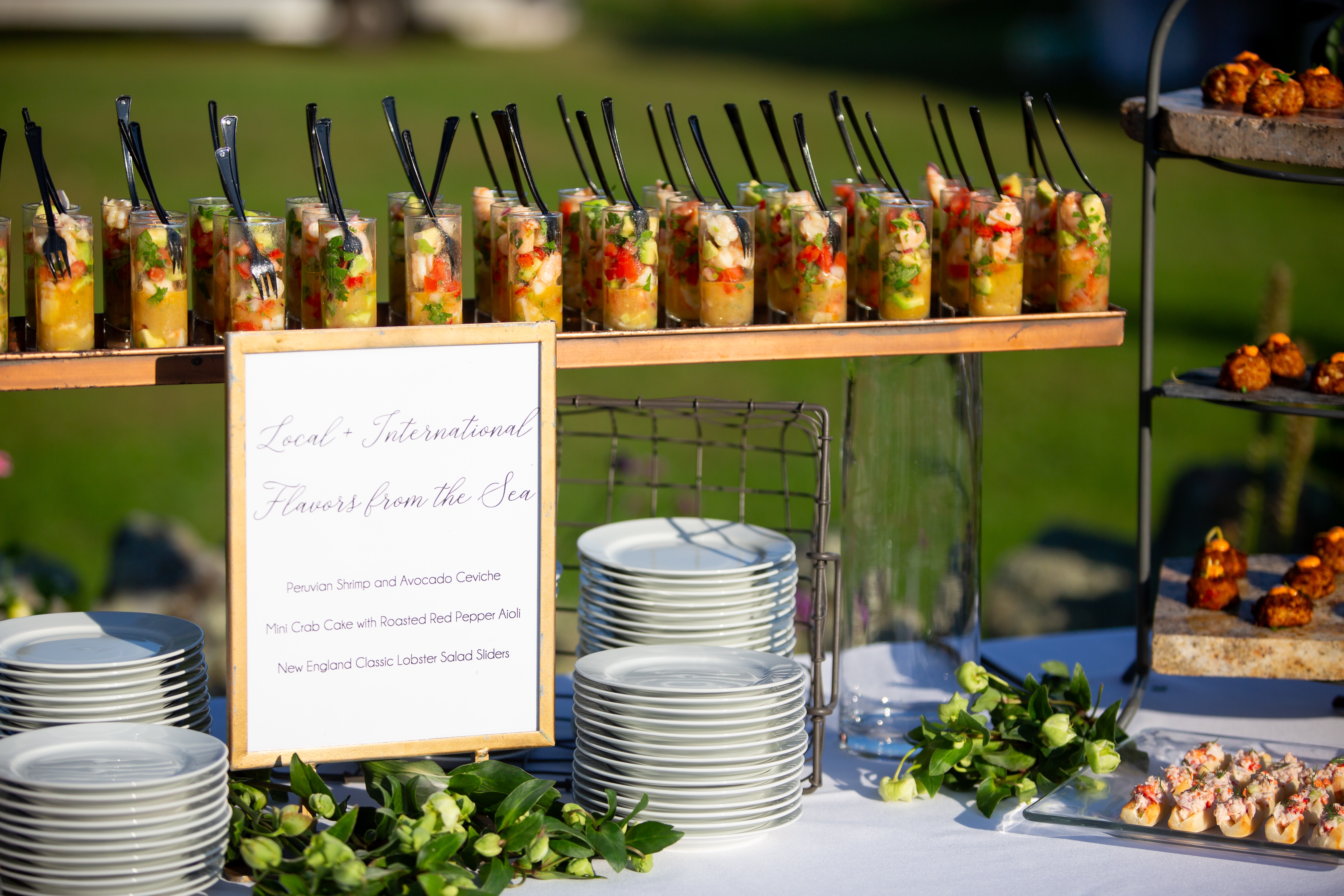 Appetizers on a table with a menu stand listing international seafood dishes surrounded by stacks of plates and fresh greenery