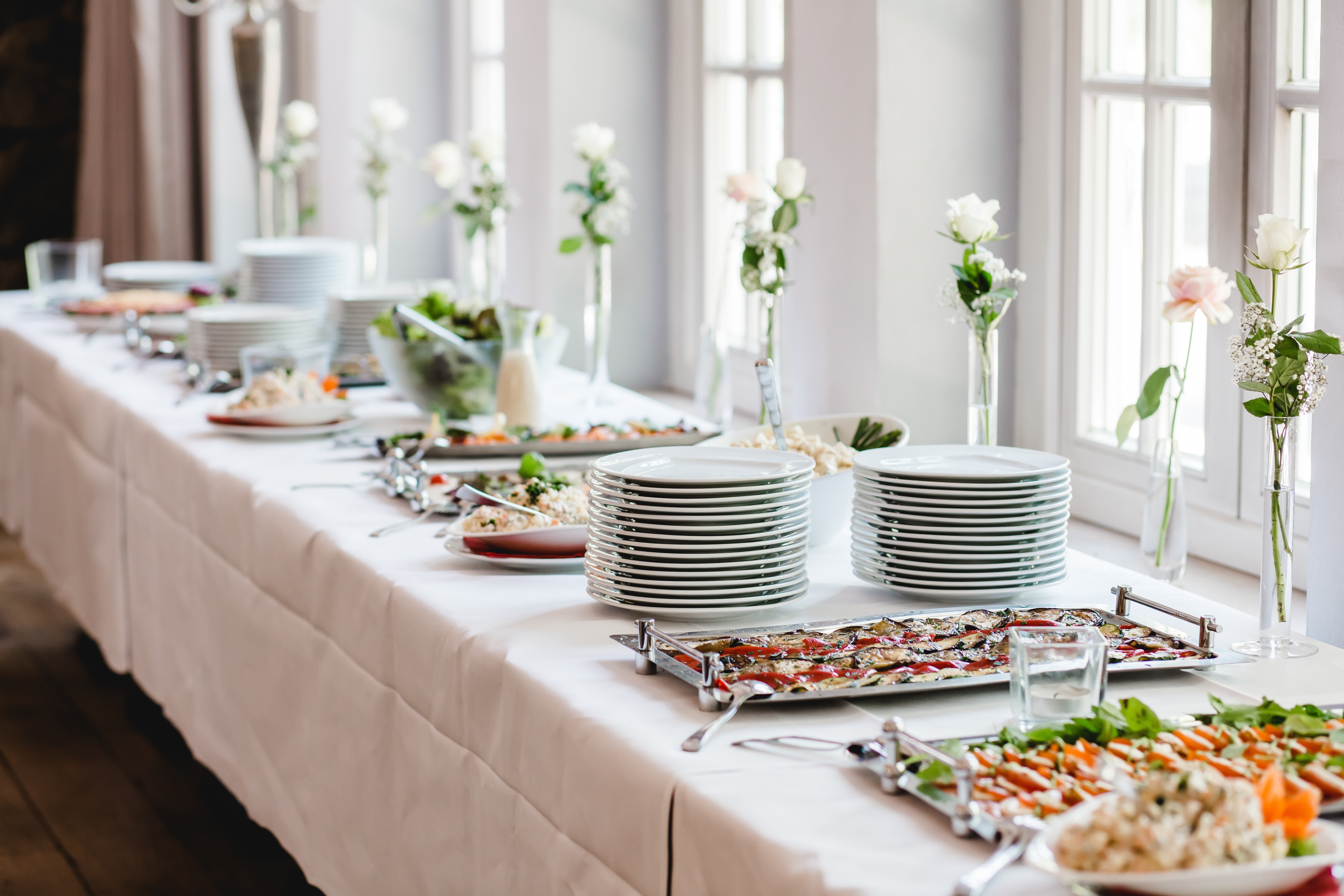 A buffet table with stacked plates, floral arrangements, and various dishes is set by a window with natural light