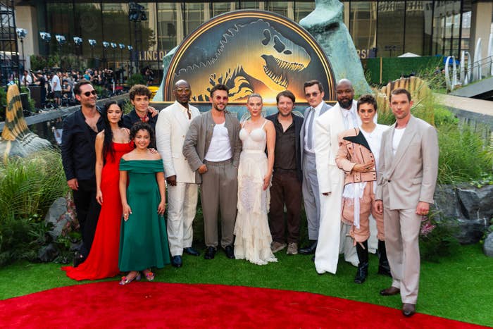 Group of actors successful ceremonial attire posing connected a reddish carpet astatine a movie premiere with a dinosaur-themed backdrop