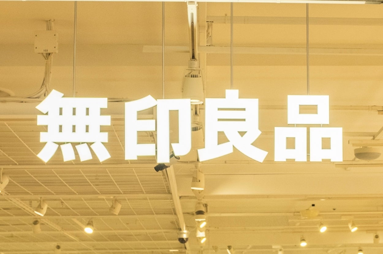 Muji store interior with shoppers browsing minimalist home goods and clothing. Illuminated sign with Japanese characters hanging from the ceiling