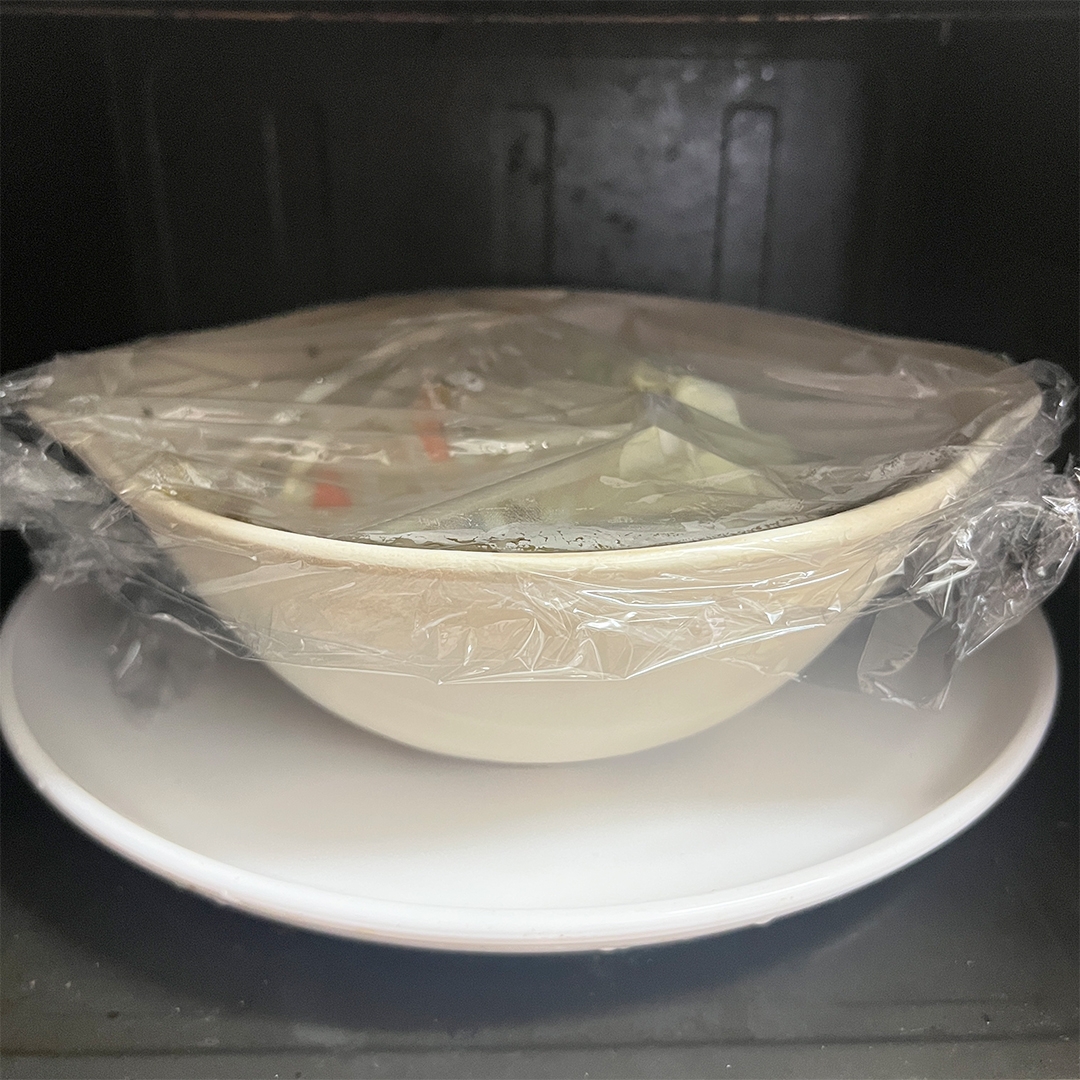 ラップをかけた白いボウルが白い皿の上に置かれており、電子レンジに入っている。