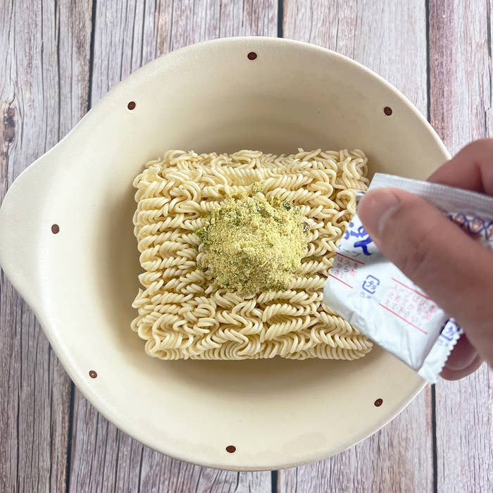 白いボウルに入った即席ラーメンの麺に、粉末スープをかけている手。