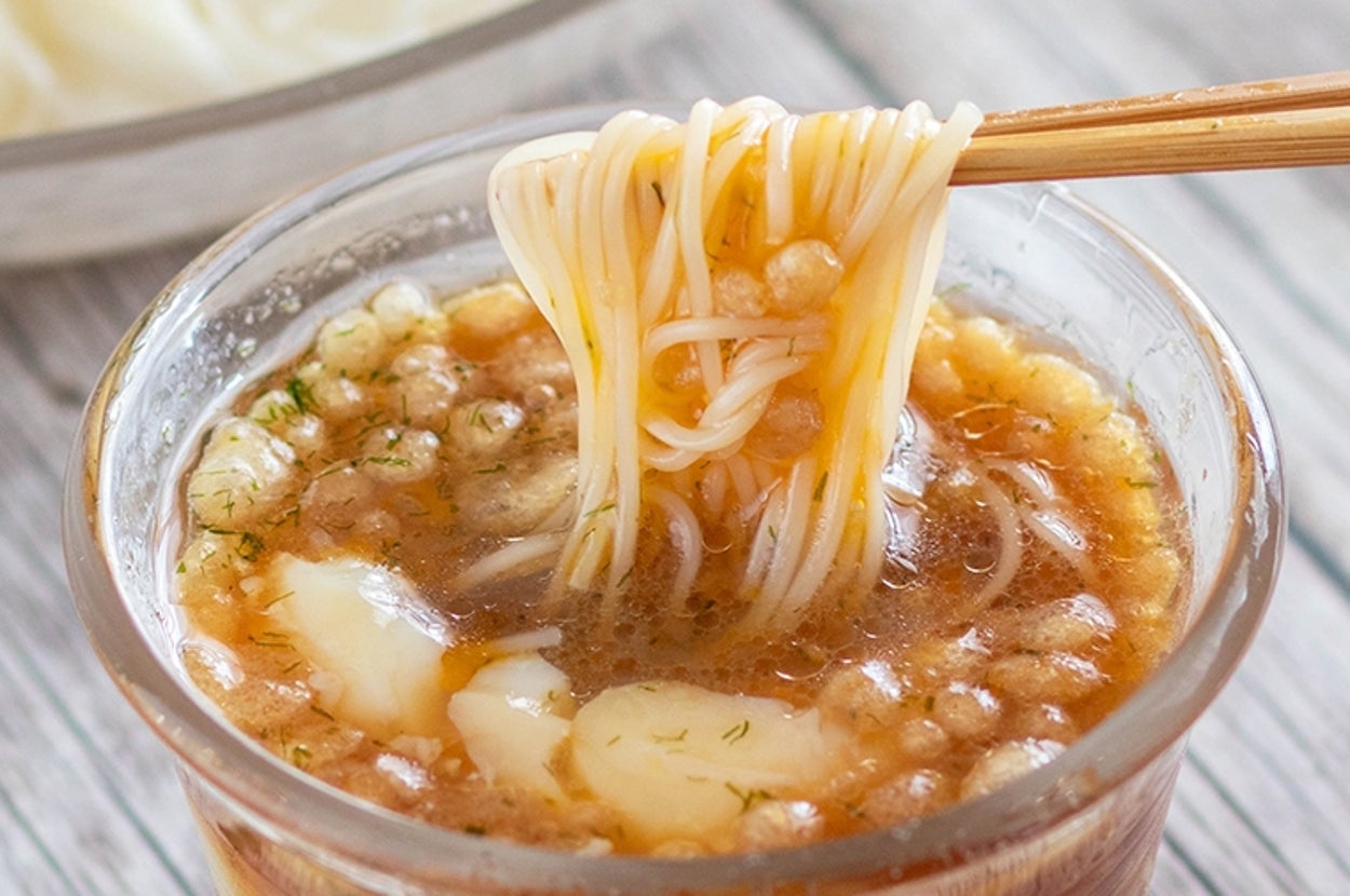 ガラスの器に入った麺つゆに箸で拾われた素麺が浸されている。背景に素麺の盛り付けがある。
