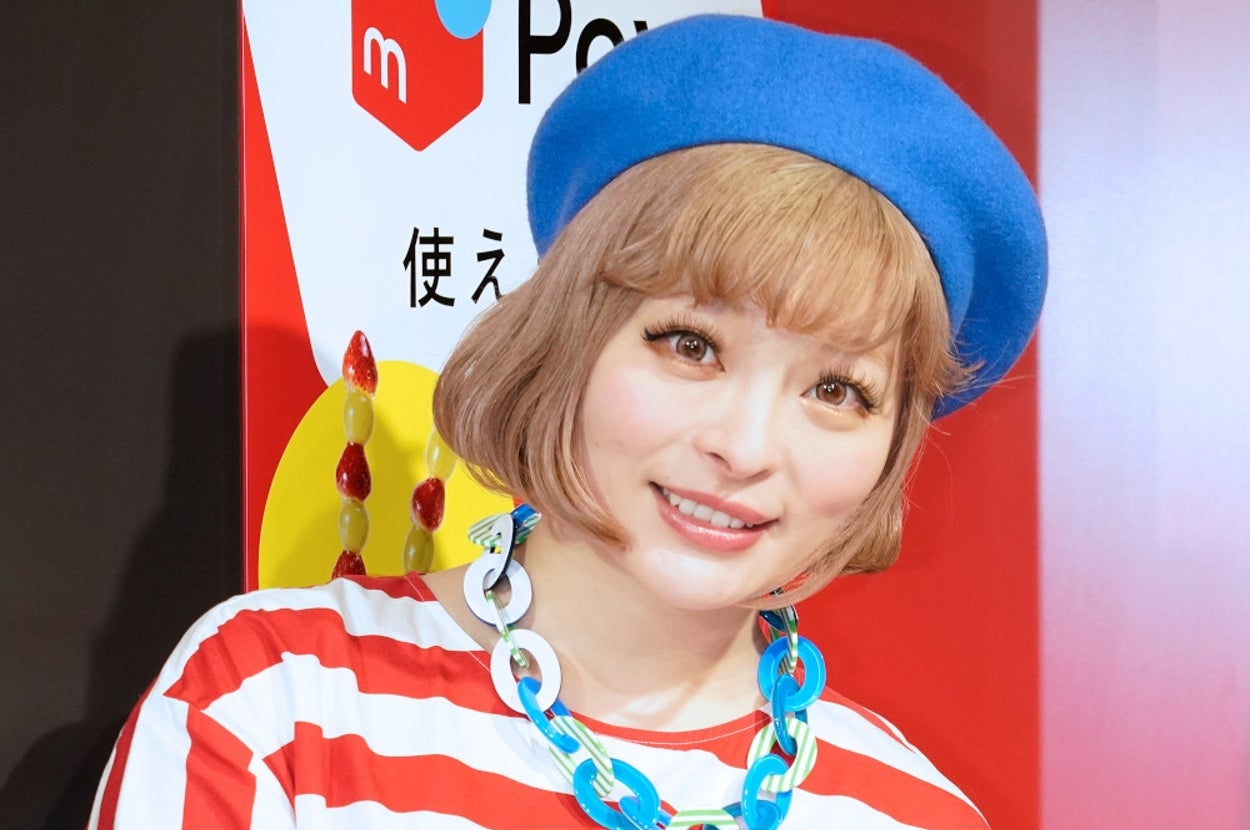 青いベレー帽とストライプの服を着た女性がポーズをとって微笑んでいます。背景に広告用の文字とロゴが見えます。
