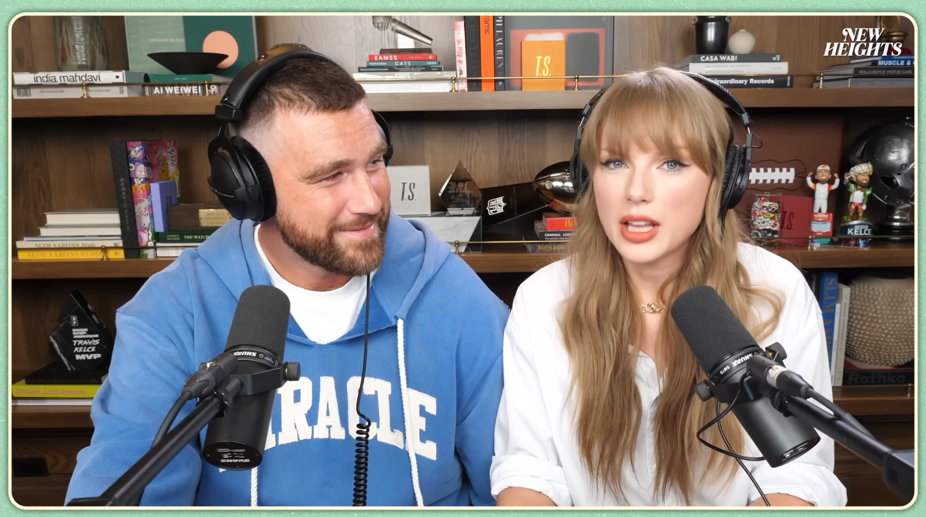 Two people wearing headsets sit in front of microphones during a podcast recording in a book-filled room