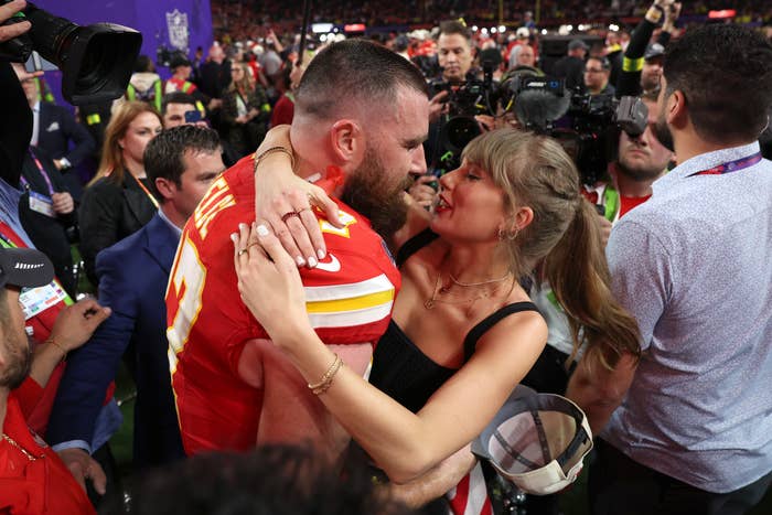 A man in a sports jersey and a woman in an elegant dress embrace surrounded by media at a sports event