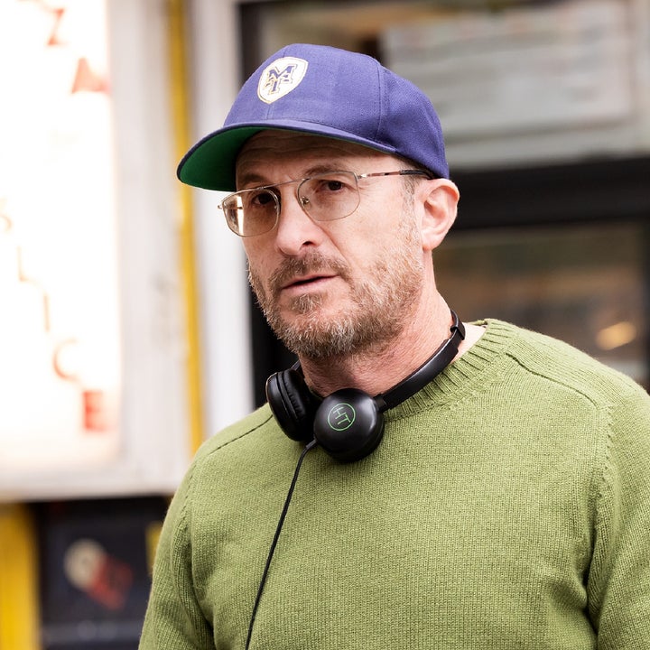 A person in casual attire with a baseball cap and headphones around their neck stands on a city street