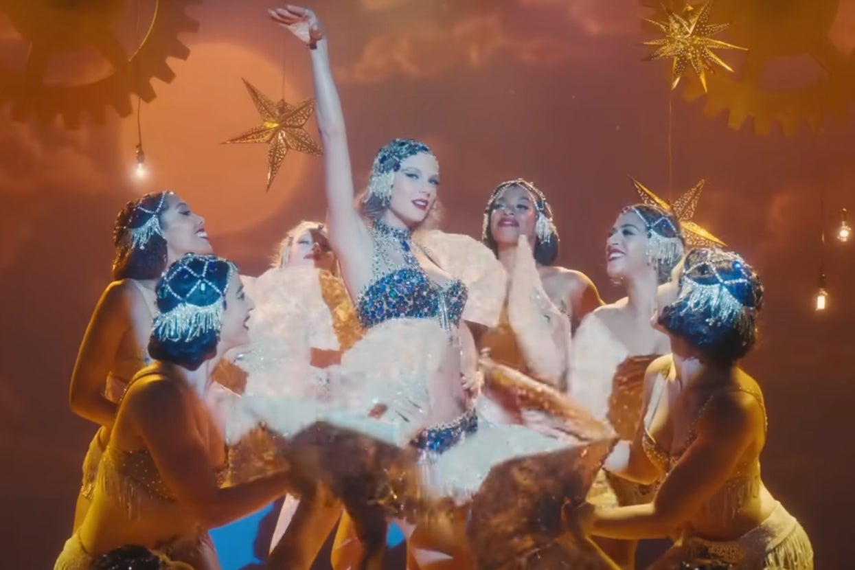 Taylor Swift in a glittering beaded costume poses elegantly surrounded by dancers with elaborate headpieces on a theatrical set with hanging stars in "Bejewled" music video