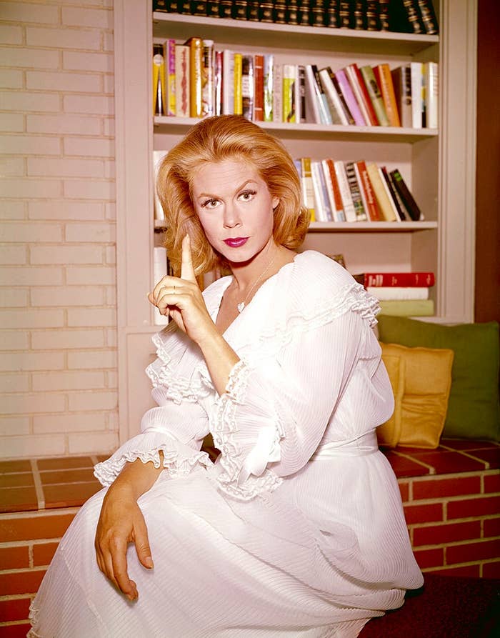 Person successful vintage, ruffled achromatic formal making a shushing gesture, seated successful beforehand of a bookshelf with assorted books displayed