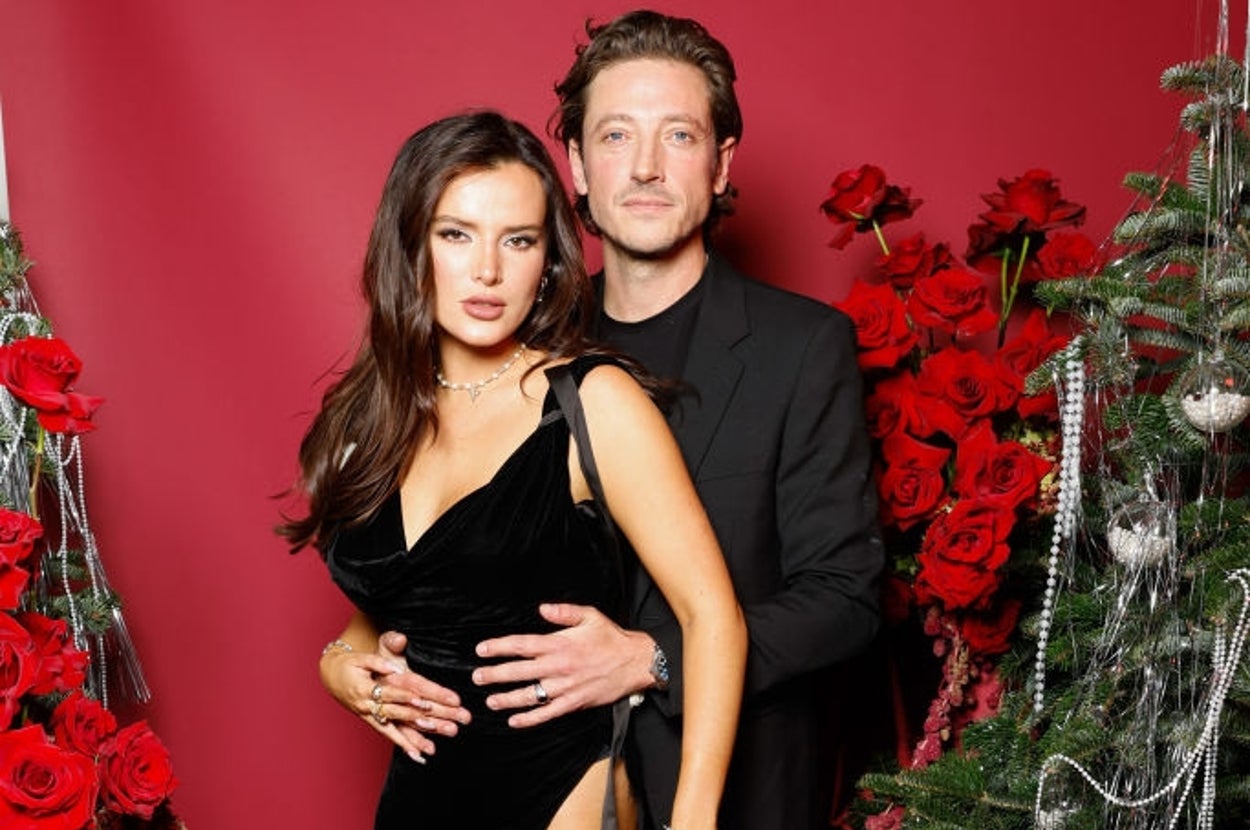 Two people pose together; one in a sleek black velvet dress with a thigh-high slit, the other in a black suit, amidst festive red and green decor