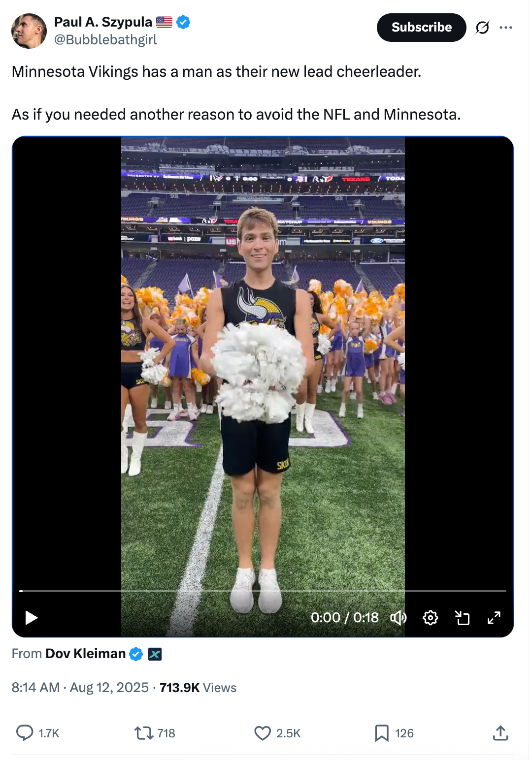 A cheerleader in uniform stands on a football field holding pom-poms, with a team of cheerleaders in the background