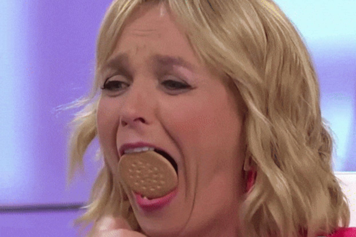A person with blonde hair attempts to fit a whole cookie into their mouth on a TV show set