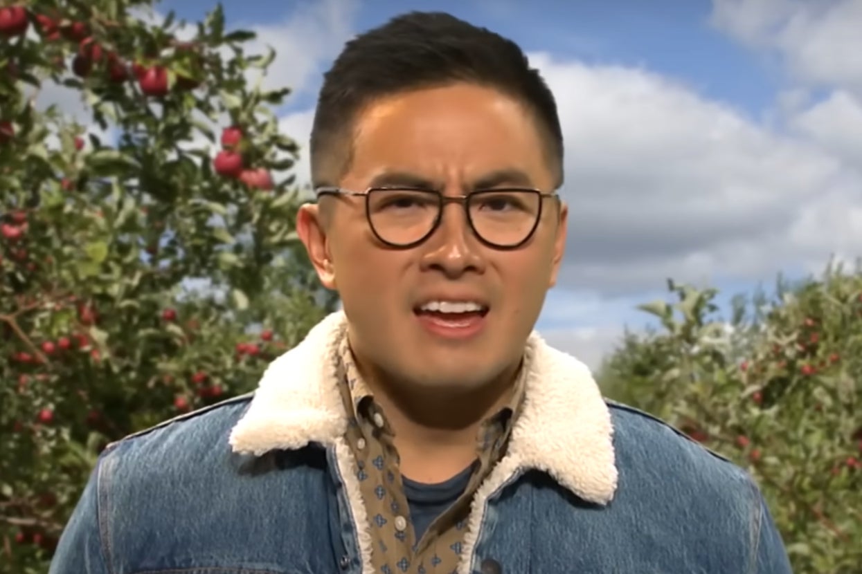 Bown Yang in and SNL skit outdoors with apple trees in the background, wearing glasses, a denim jacket with shearling collar, and a patterned shirt