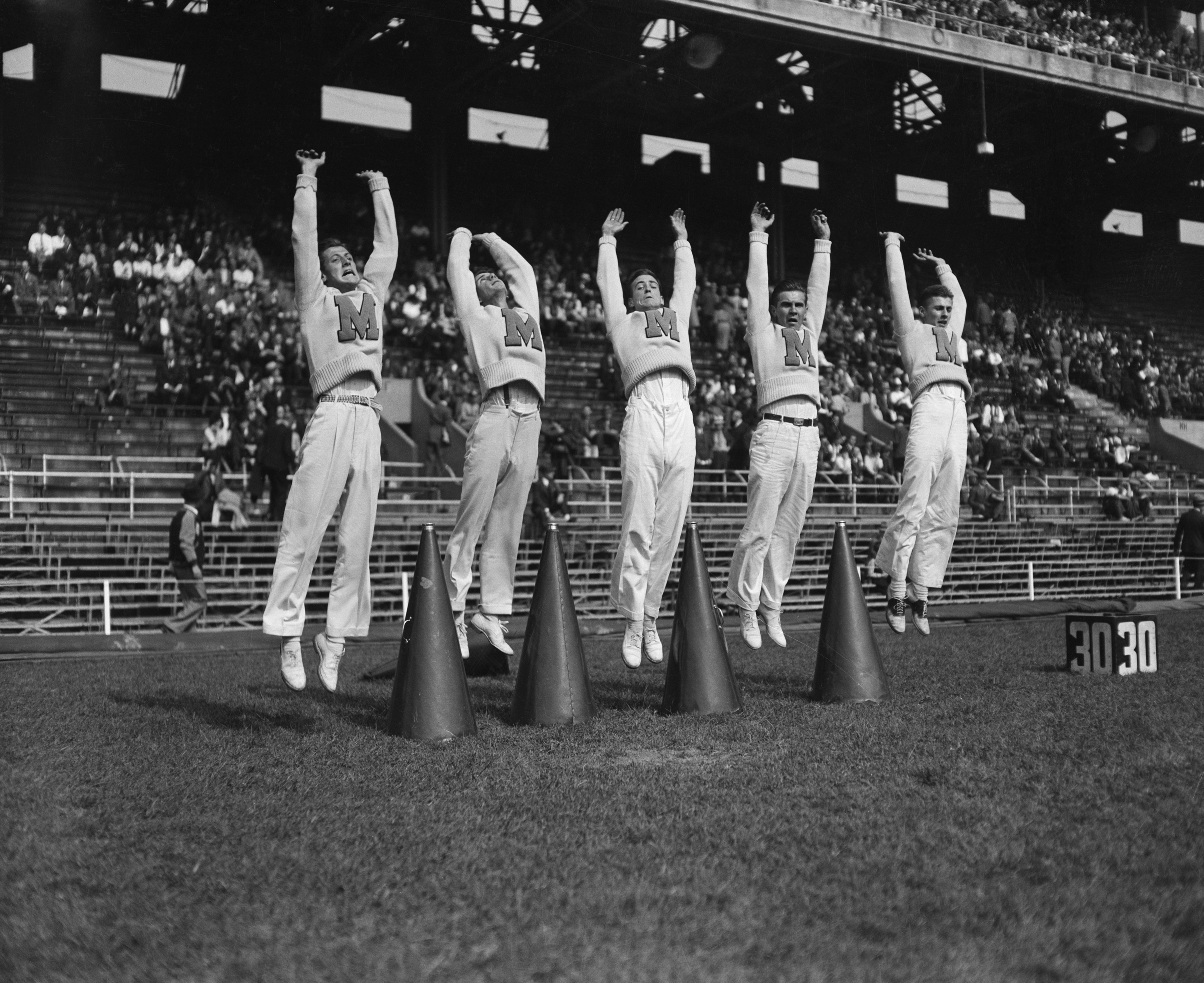 Five athletes in identical athletic outfits jump in unison at a track field event, with numbered markers on the ground