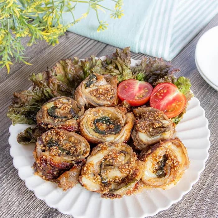 皿に盛られた肉巻き野菜の料理。ゴマがトッピングされ、トマトと葉野菜が添えられている。