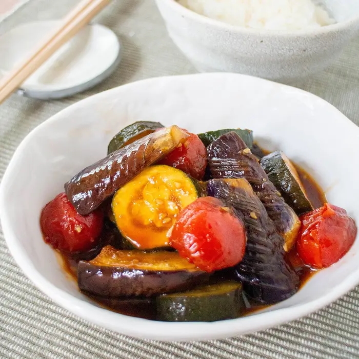 皿に盛られた焼き野菜とトマトの料理。後ろにご飯と箸が置かれている。