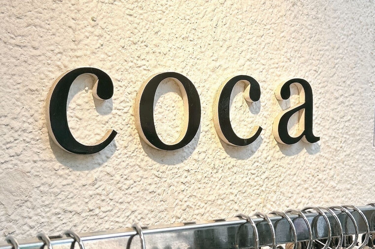 洋服店の壁に「coca」の文字が飾られている。下には服が掛けられたラックが見える。