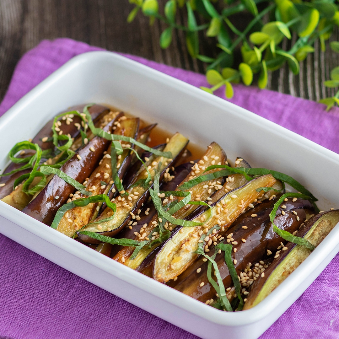 白い皿に盛られた焼きナス、ゴマと細切りの青じそがトッピングされている。背景に観葉植物が見える。