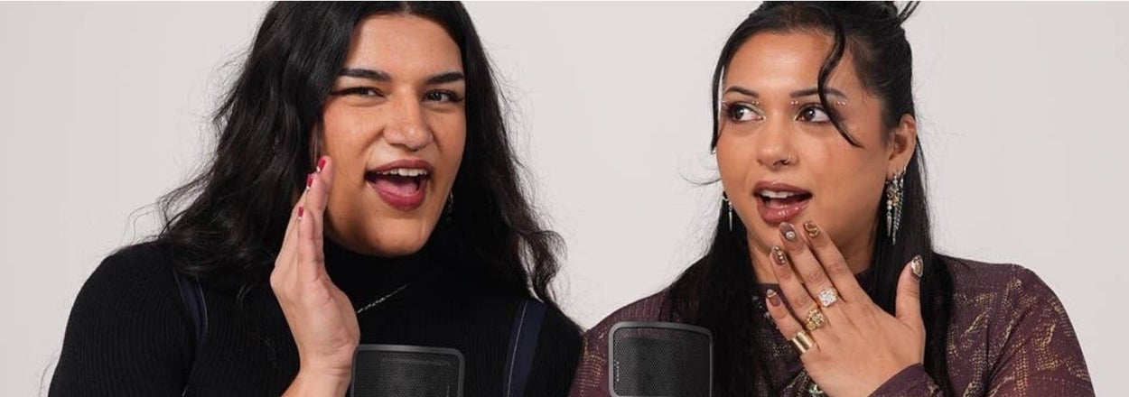 Two people sitting, holding microphones, laughing and chatting. One wears a long-sleeve top and pants; the other wears an overalls-style outfit
