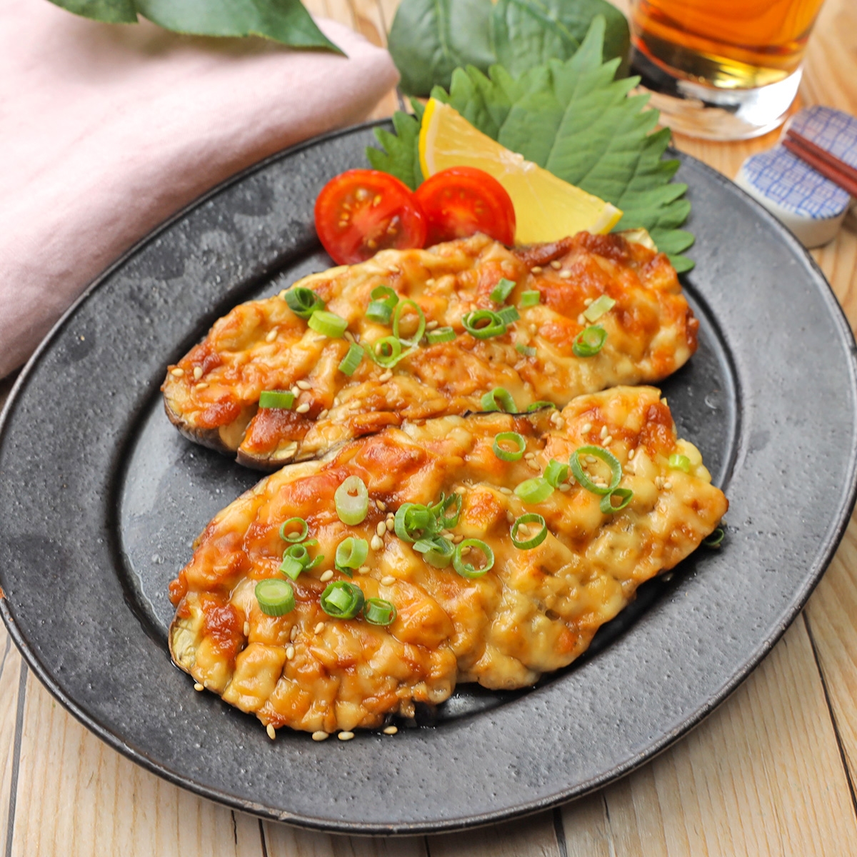 焼きナスにみそのタレがかかり、ネギとトマト、レモンが添えられた料理。木のテーブルに置かれている。
